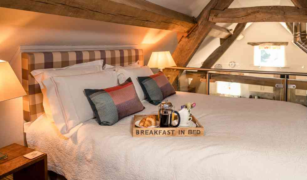 Cosy bedroom at Cruck Barn Cottage with checked headboard, crisp white bedding, and breakfast in bed tray with coffee and croissants.